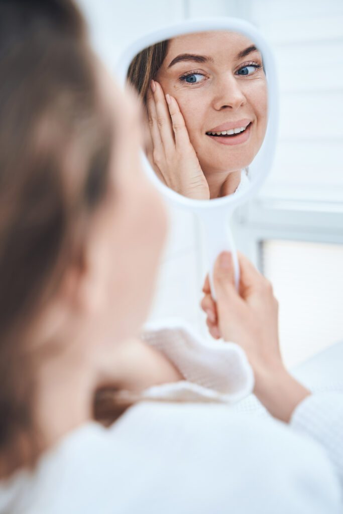 Kind female person holding mirror in her hand while touching face after procedure